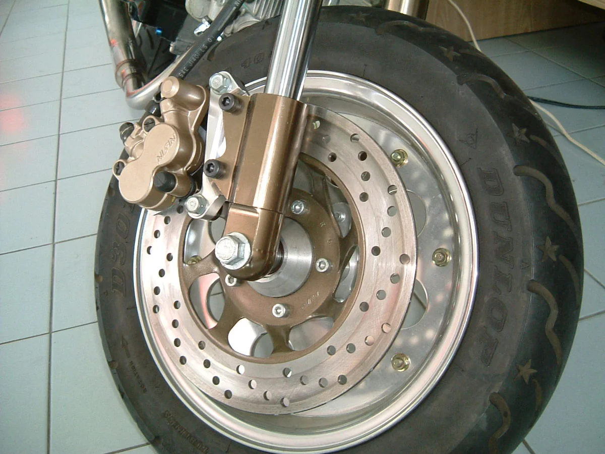 A motorcycle front wheel with a drilled stainless brake disc, hub and caliper mounted to the fork leg.