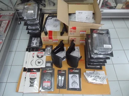 Motorcycle parts laid out on a work mat with their printed packaging and part-number labels: levers, footpegs, mirrors, brackets and small fittings.