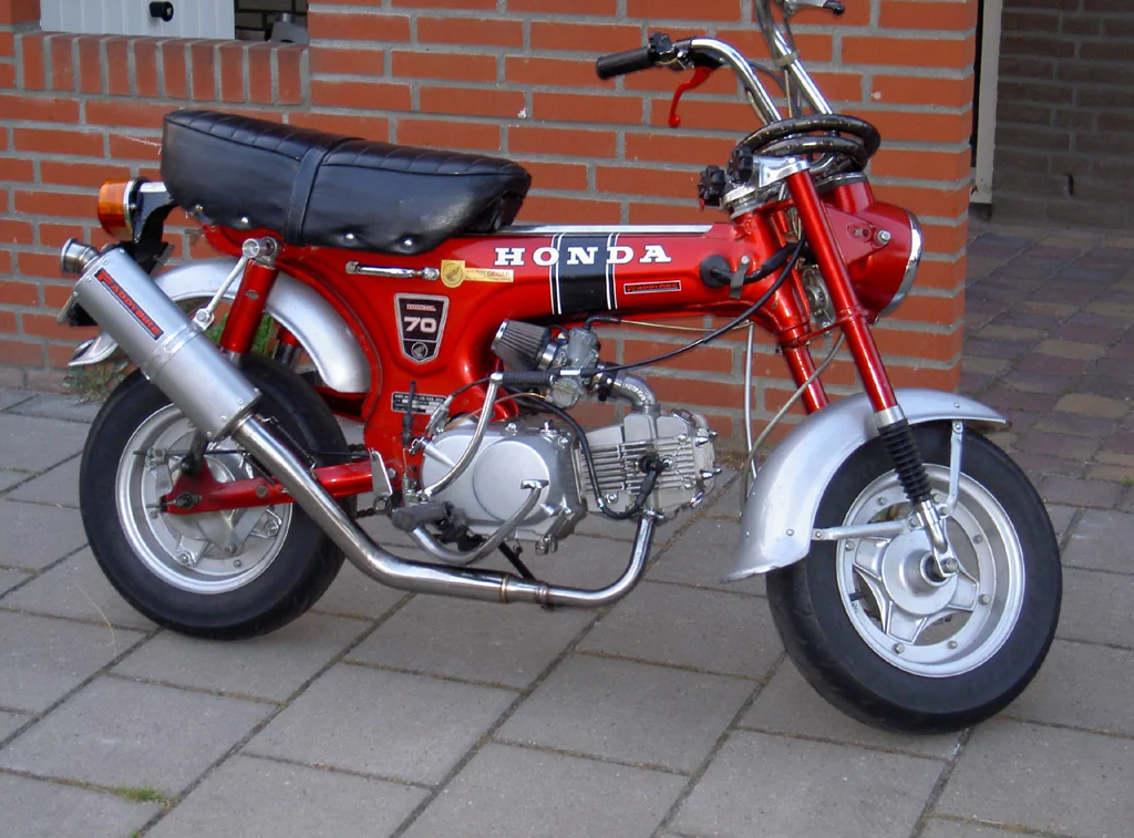 A red Honda Dax mini motorcycle with chrome exhaust and a black seat, parked on paving.